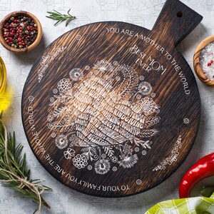 May include: A round wooden cutting board with a burnt finish. The board has a white design of flowers and words that say "To your family you are the whole world. You are a mother to the world. Best Mom Ever."