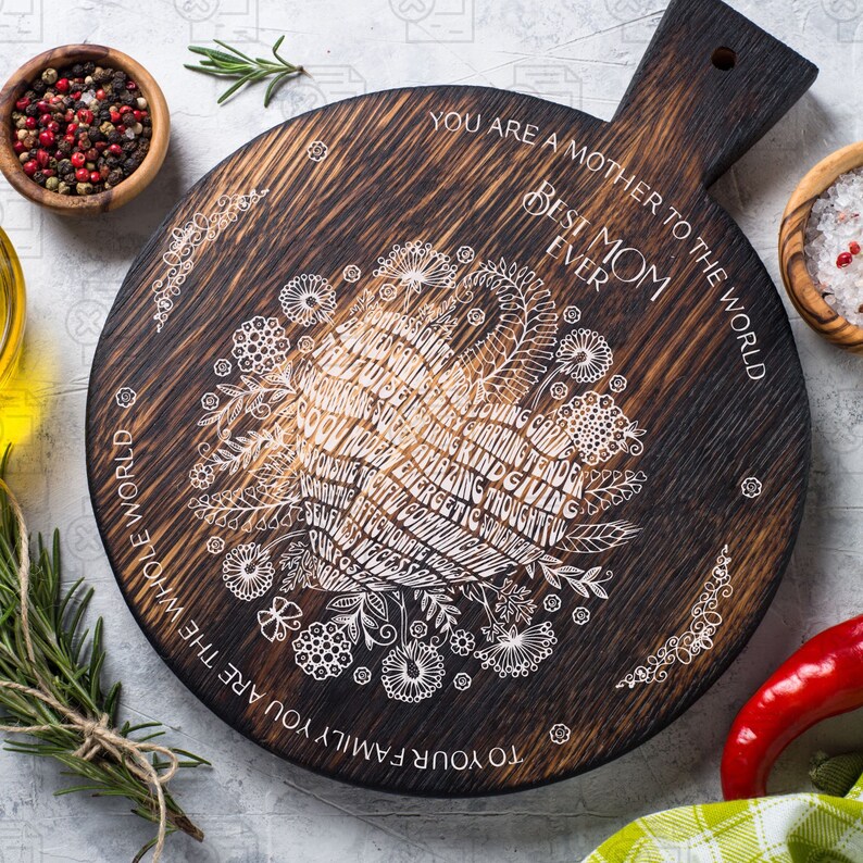 May include: A round wooden cutting board with a burnt finish. The board has a white design of flowers and words that say "To your family you are the whole world. You are a mother to the world. Best Mom Ever."