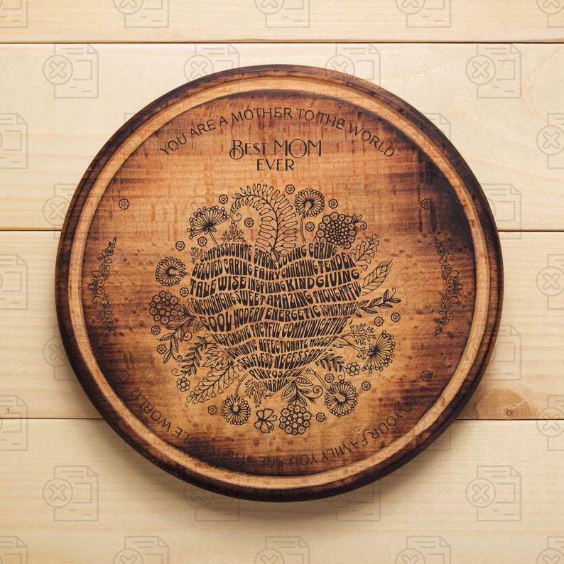 May include: A round wooden cutting board with a heart-shaped design in the center. The heart is filled with words describing a mother, such as "loving", "caring", "kind", and "amazing". The text "You are a mother to the world Best Mom Ever" is written around the heart.