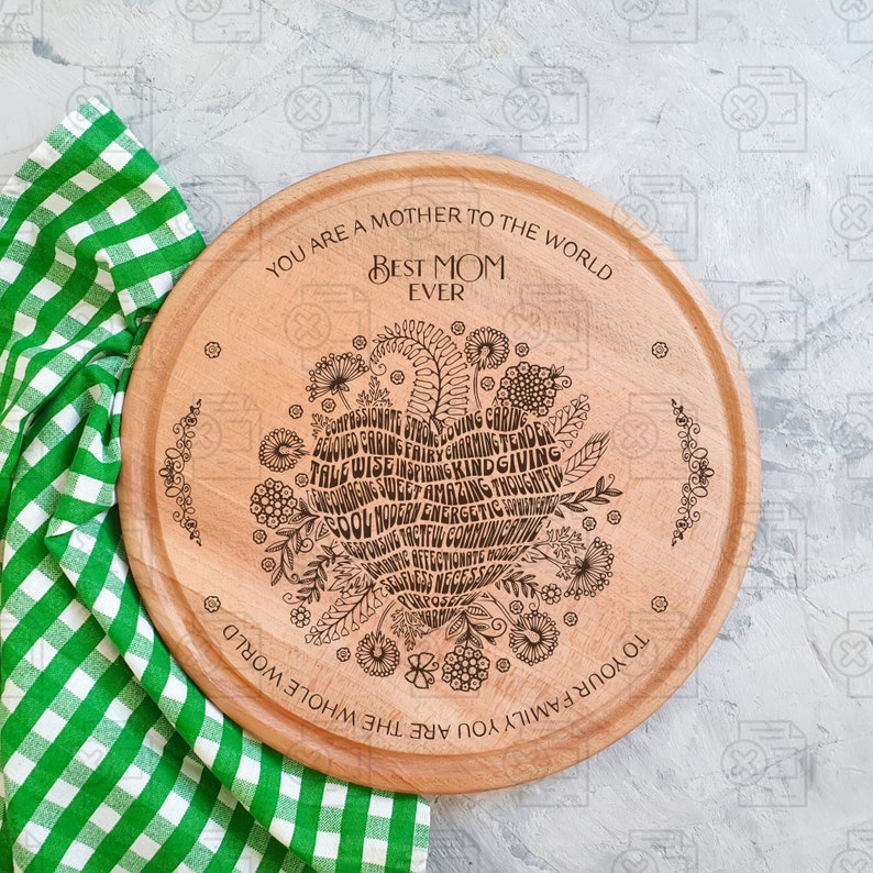 May include: A round wooden serving board with a laser-engraved design. The design features a heart shape filled with words describing positive qualities, such as "compassionate", "loving", "caring", "wise", "inspiring", "kind", and "giving". The text "You are a mother to the whole world. Best Mom Ever. To your family you are the whole world" is engraved around the edge of the board.