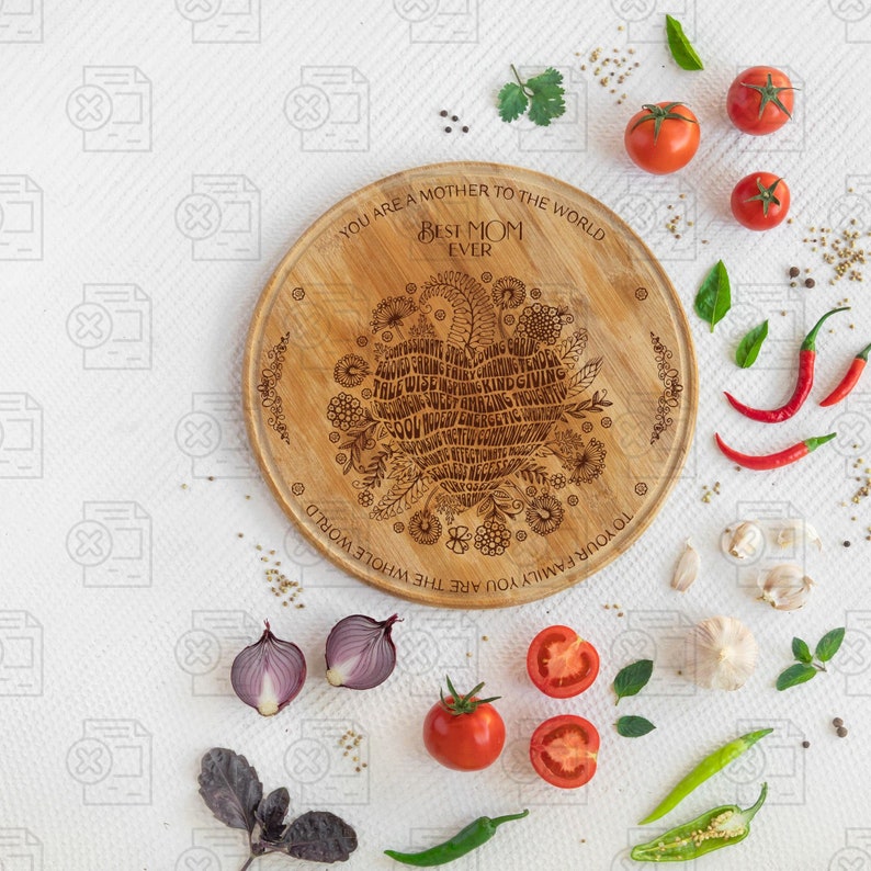 May include: A round wooden serving board with a heart-shaped design engraved with the words "You are a mother to the whole world. To your family you are the whole world. Best Mom Ever." The board is surrounded by various vegetables and herbs, including tomatoes, onions, garlic, and green peppers.