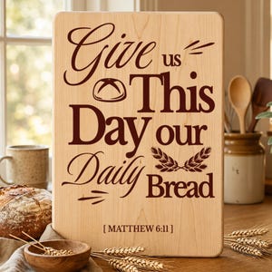 May include: A rectangular light-colored wooden cutting board featuring dark brown engraved text that reads Give us This Day our Daily Bread with a small bread loaf icon, wheat stalks, and the citation Matthew 6:11 at the bottom center.