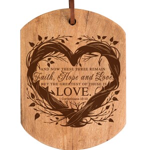 Puede incluir: Un letrero de madera en forma de coraz&oacute;n con un acabado de madera marr&oacute;n. El letrero presenta un dise&ntilde;o de rama de &aacute;rbol estilizado con el texto "AND NOW THESE THREE REMAIN Faith, Hope and Love BUT THE GREATEST OF THESE IS LOVE. 1 Corinthians 13:13".