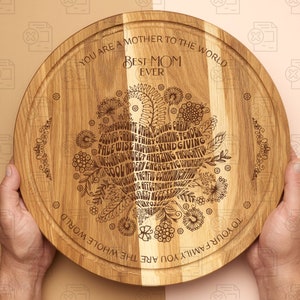 May include: A round wooden serving board with a heart-shaped design engraved with the words "You are a mother to the whole world. Best Mom Ever." and a list of positive adjectives. The board is made of light brown wood with a natural grain.