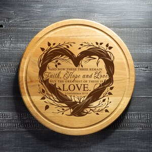 Puede incluir: Una tabla de cortar redonda de madera con un dise&ntilde;o en forma de coraz&oacute;n grabado con el texto "AND NOW THESE THREE REMAIN Faith, Hope and Love. BUT THE GREATEST OF THESE IS LOVE. 1 Corinthians 13:13."