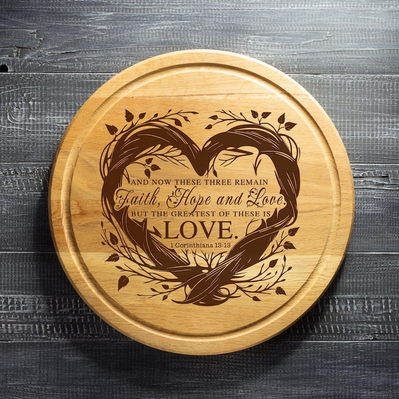 Puede incluir: Una tabla de cortar redonda de madera con un dise&ntilde;o en forma de coraz&oacute;n grabado con el texto "AND NOW THESE THREE REMAIN Faith, Hope and Love. BUT THE GREATEST OF THESE IS LOVE. 1 Corinthians 13:13."