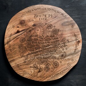 May include: A round wooden serving tray with a laser-engraved design. The design features a heart shape with the words "Best Mom Ever" and a circle of words describing positive qualities. The words are "You are a mother to the world" and "To your family you are the whole world" are also engraved on the tray.