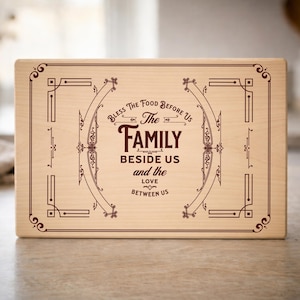 Pode incluir: Uma tábua de corte retangular de madeira clara com uma borda gravada em marrom escuro e o texto: Bless The Food Before Us The FAMILY BESIDE US and the LOVE BETWEEN US, centralizado dentro de um design de moldura decorativa.