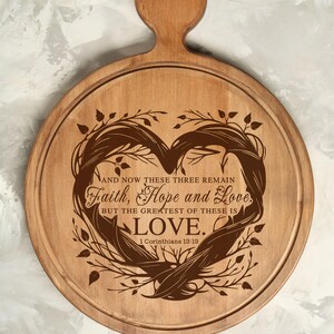 Puede incluir: Una tabla de cortar redonda de madera con un dise&ntilde;o en forma de coraz&oacute;n y el texto "AND NOW THESE THREE REMAIN Faith, Hope and Love. BUT THE GREATEST OF THESE IS LOVE. 1 Corinthians 13:13" grabado en ella.