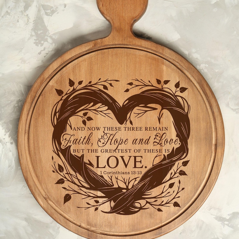 Puede incluir: Una tabla de cortar redonda de madera con un dise&ntilde;o en forma de coraz&oacute;n y el texto "AND NOW THESE THREE REMAIN Faith, Hope and Love. BUT THE GREATEST OF THESE IS LOVE. 1 Corinthians 13:13" grabado en ella.