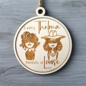 May include: A round wooden ornament with a laser-cut design of two cartoon figures wearing sunglasses. The text "Every Thelma Needs a Louise" is written around the figures.