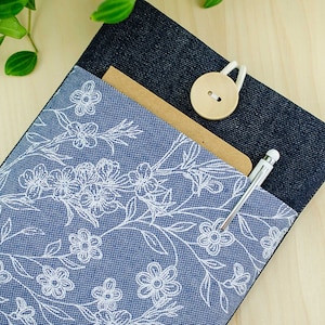 May include: A blue denim fabric pouch with a white floral pattern. The pouch has a brown paper pocket and a wooden button closure. A silver pen is sticking out of the pocket.
