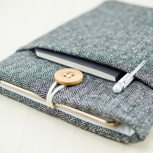 May include: A gray and white herringbone fabric tablet sleeve with a black notebook and a silver pen inside. A wooden button is attached to the sleeve with a white string.