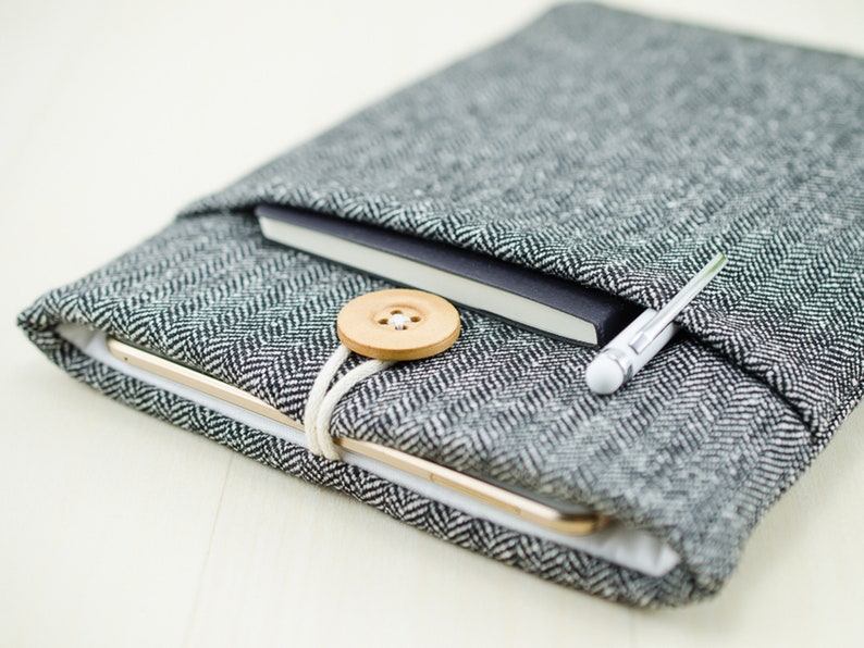 May include: A gray and white herringbone fabric tablet sleeve with a black notebook and a silver pen inside. A wooden button is attached to the sleeve with a white string.