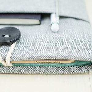 May include: A gray fabric case with a black button closure, a white pen, and a tablet computer inside. The case has a light green lining.