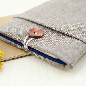 May include: A brown linen tablet sleeve with a wooden button closure and a blue lining. The sleeve is folded over and resting on a brown surface.