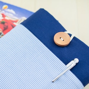 May include: A blue and white striped fabric pouch with a blue fabric flap. The pouch has a wooden button closure and a silver pen tucked into the flap.