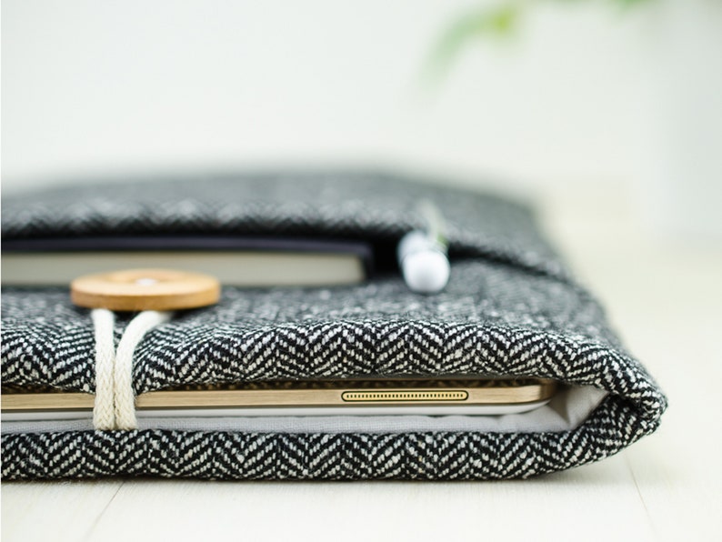 May include: A black and white herringbone fabric laptop sleeve with a wooden button and a white cord. The sleeve is closed and a gold tablet is visible inside.