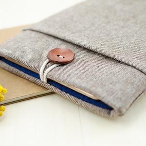Könnte beinhalten: Eine braune Tablet-Hülle aus Leinen mit einem Holz-Knopfverschluss und einem blauen Futter. Die Hülle ist umgeklappt und liegt auf einem braunen Notizbuch.