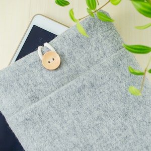 May include: A gray felt tablet sleeve with a wooden button closure. The sleeve is lying on a wooden surface with a tablet inside.