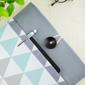 Könnte beinhalten: Eine graue Stoff-Notizbuchhülle mit einem geometrischen Muster aus weißen, rosa und türkisfarbenen Dreiecken. Ein silberner Stift befindet sich in der Tasche, und ein dunkelbrauner Knopf sichert die Hülle. Die Notizbuchhülle liegt auf einer hellen Holzoberfläche.