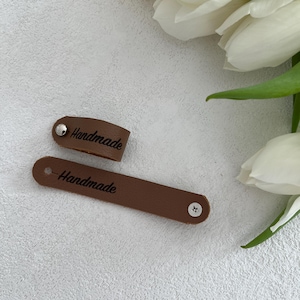 Könnte beinhalten: Zwei braune Lederanhänger mit dem Aufdruck "Handmade". Einer ist eine kleine Schlaufe, der andere ein langer, dünner Streifen. Beide Anhänger haben eine silberne Niete. Die Anhänger liegen auf einer strukturierten, hellgrauen Oberfläche.