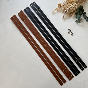 Puede incluir: Varias correas de cuero en negro y marrón, cada una con remaches de color latón. Las correas están dispuestas en diagonal sobre una superficie blanca, con flores secas en el fondo. Las correas de cuero son para un proyecto artesanal.