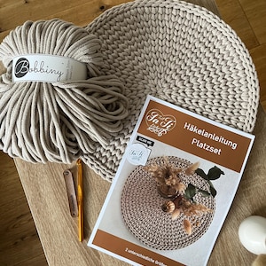 Könnte beinhalten: Ein beiger gehäkelter Platzset mit passendem Bobbiny-Garn, einer Häkelnadel und einem Anleitungsheft. Das Heft trägt die Aufschrift "Häkelanleitung Platzset" und "2 unterschiedliche Größen".