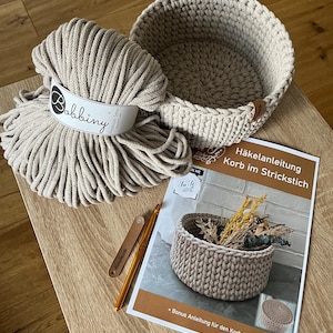 Pode incluir: Uma cesta de crochê bege com um livreto de instruções de crochê, uma bola de corda de algodão bege e um gancho de crochê dourado. O livreto tem o texto "Häkelanleitung Korb im Strickstich" e "Bonus Anleitung für den Korbdeckel".