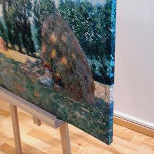 以下が含まれることがあります： 田園風景の印象派絵画。大きな干し草の山が野原に描かれています。緑、黄、青色のパレットで、背景には木々があります。キャンバスは木製のイーゼルに展示されています。