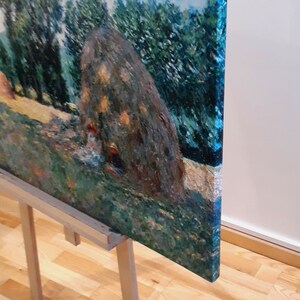 May include: A painting on a wooden easel depicts a landscape with a large haystack in the foreground. The haystack is painted in shades of brown and green, with a few highlights of yellow. The background is a lush green field with a row of trees in the distance. The painting is framed in a simple wooden frame.