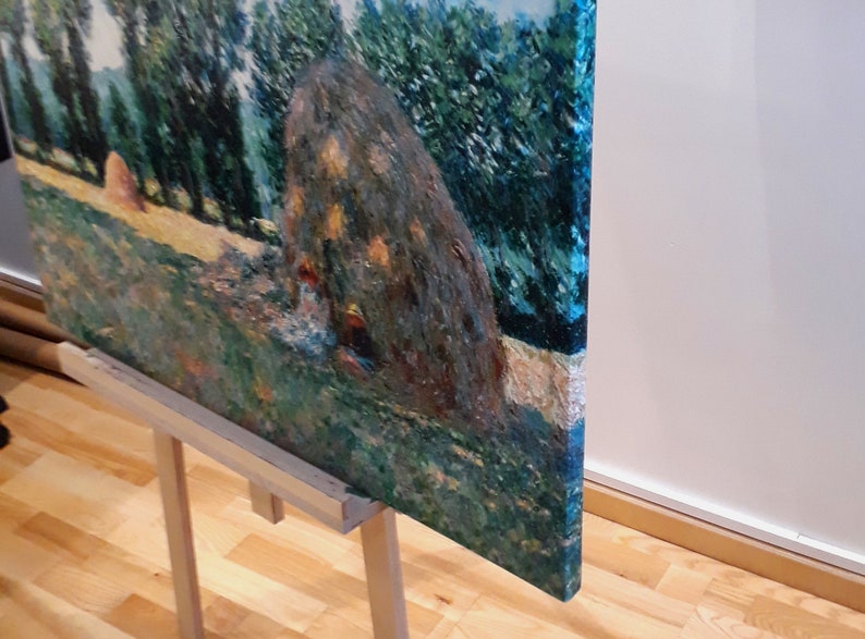May include: A painting on a wooden easel depicts a landscape with a large haystack in the foreground. The haystack is painted in shades of brown and green, with a few highlights of yellow. The background is a lush green field with a row of trees in the distance. The painting is framed in a simple wooden frame.