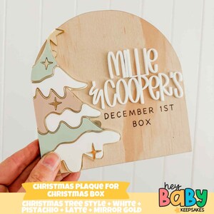 May include: A wooden Christmas plaque with a Christmas tree design in white, pistachio, and latte, with gold mirror accents. It displays the names "Millie & Cooper's" and the date "December 1st". A festive holiday decoration.