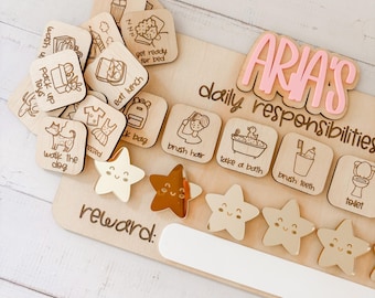 Puede incluir: Un cuadro de madera con el nombre "ARIA'S daily responsibilities" en la parte superior. El cuadro tiene una serie de fichas de madera con imágenes y texto, como "brush hair", "take a bath", "brush teeth" y "toilet". También hay estrellas de madera con caras sonrientes que se pueden usar como recompensas.