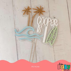 May include: A white acrylic cake topper with the text "Cooper is 1" in white, green, and brown. The topper features palm trees, a wave, and a surfboard.