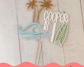Puede incluir: Un topper de pastel de acrílico blanco con el texto "Cooper is 1" en blanco, verde y marrón. El topper presenta palmeras, una ola y una tabla de surf.