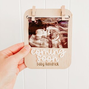 Op de afbeelding: Een houten fotolijst met een witte achtergrond en een zwart-witte echografie afbeelding. De lijst heeft een clip aan de bovenkant om de afbeelding vast te houden. De tekst "coming soon baby kendrick" staat op de lijst.