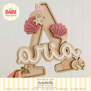May include: A wooden letter A plaque featuring the name Aria in white script. The design is decorated with dusty rose acrylic seashells and mirror gold starfish accents. The top left corner includes a circular logo reading hey BABY keepsakes.