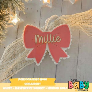 May include: A personalised bow-shaped Christmas ornament in raspberry sorbet with a white scalloped edge and gold mirror text reading "Millie". It has a white rope for hanging. The text "Personalised Bow Ornament" is at the bottom.