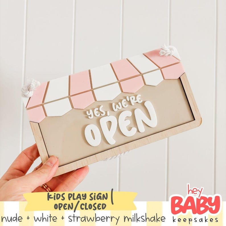 May include: A rectangular wooden sign featuring a light beige background with the white raised text "YES, WE'RE OPEN". The top of the sign has a decorative awning pattern in alternating light pink and white segments, with white rope loops at the corners.