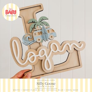 Op de afbeelding: Een houten letter L-bord met een gelaagd ontwerp van een lichtblauwe camper, een surfplank, een saliegroene palmboom en een lichtblauwe golf. De naam Logan is in witte cursieve letters in het midden van de letter aangebracht.
