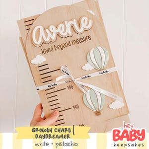 Può includere: Un metro di crescita in legno chiaro con parte superiore ad arco, il nome Averie e il testo loved beyond measure. Include segni di misurazione neri, due sagome di mongolfiere verde pallido e nuvole bianche, legato con un nastro bianco.