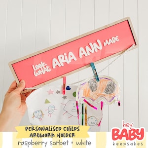 May include: A personalized child's artwork holder with the text "ARIA ANN MADE" in white letters on a raspberry sorbet background. The wooden frame holds children's drawings with clothespins. The artwork holder is labeled "hey BABY keepsakes."