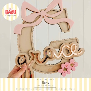 May include: A wooden letter G wall plaque featuring a light pink bow at the top and two light pink flowers at the bottom. The name grace is written in a rose gold mirror script font across the centre of the letter. The edges have a scalloped design.