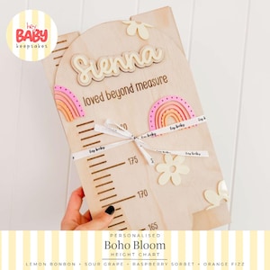 May include: A light-colored wooden height chart with an arched top, featuring the name Sienna in raised script, the text loved beyond measure, and decorative wooden flowers and rainbows in shades of pink and orange, tied with a branded ribbon.