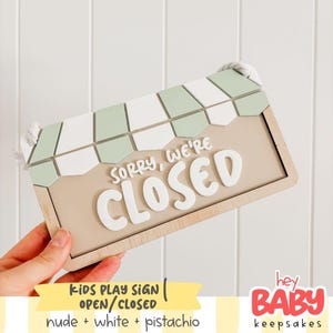 May include: A rectangular wooden sign featuring a light beige background with the white raised text "SORRY, WE'RE CLOSED". The top of the sign has a decorative awning pattern in alternating sage green and white segments, with white rope attached.