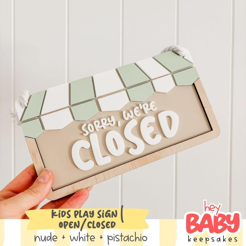 May include: A rectangular wooden sign featuring a light beige background with the white raised text "SORRY, WE'RE CLOSED". The top of the sign has a decorative awning pattern in alternating sage green and white segments, with white rope attached.