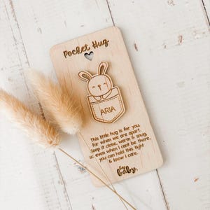 May include: A wooden pocket hug with a laser-cut bunny in a pocket design. The text "Pocket Hug" and "Aria" are visible. The card includes a message about keeping the hug close. Dried flowers are in the background.