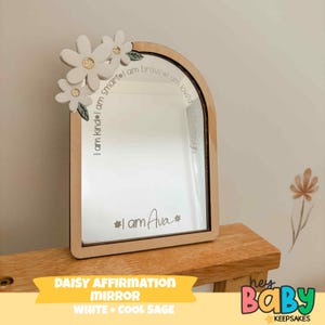 May include: Wooden framed mirror with a daisy flower design. The mirror has the text "I am kind, I am smart, I am brave, I am loved, I am enough" and "I am Ava". The mirror is labeled "DAISY AFFIRMATION MIRROR WHITE + COOL SAGE".