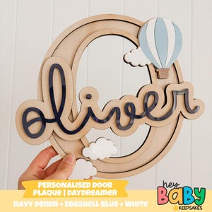 Puede incluir: Placa de puerta de madera personalizada con el nombre "oliver" en escritura azul marino. La placa presenta un globo aerostático azul claro y blanco y nubes blancas. El texto "PERSONALISED DOOR PLAQUE | DAYDREAMER" está en la parte inferior.